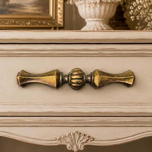 May include: A horizontal drawer pull handle with a metallic antique brass finish, featuring a central ribbed sphere flanked by two elongated, tapered arms with faceted edges, mounted on a light beige, distressed wooden drawer front.