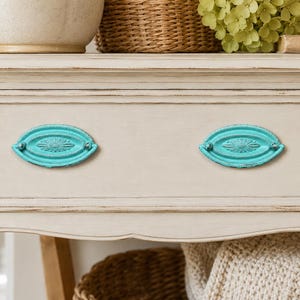 May include: A light beige wooden drawer front featuring two oval-shaped turquoise metal pulls. Each pull has a decorative embossed sunburst pattern in the center and a distressed finish, mounted horizontally on the flat surface of the drawer.