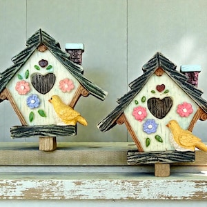 May include: Two decorative birdhouses featuring a cream-colored facade with a dark brown heart-shaped opening, a yellow bird figure, and relief-molded pink and blue flowers. Each house has a dark green textured roof and a small chimney.