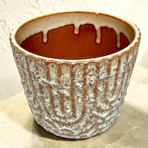May include: A ceramic planter with a textured, ridged exterior in shades of brown and white. The rim features a dripping glaze effect, and the interior is a warm brown color. This decorative pot is suitable for indoor plants.