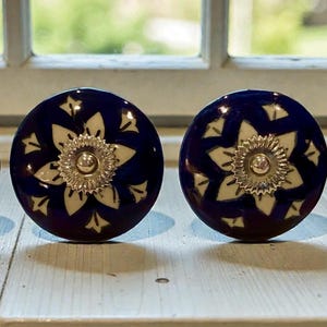 May include: Four round cabinet knobs featuring a deep blue background with a white floral pattern and a central silver-toned metal rosette. Each knob has a circular shape with a smooth finish, showcasing a repeating petal design radiating from the center.