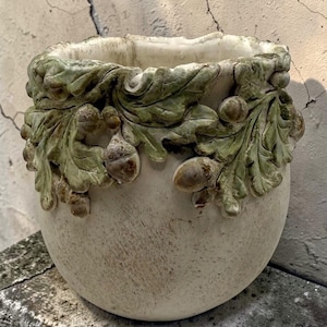 Puede incluir: Una maceta redonda de color blanquecino con una superficie texturizada. El borde superior presenta un borde decorativo de hojas verdes y bellotas marrones. Esta maceta es adecuada para uso en interiores o exteriores.