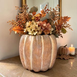 May include: A round, ribbed ceramic vase with a weathered, light brown finish shaped like a pumpkin. The vase is filled with an arrangement of artificial autumn foliage, including dried oak leaves, small orange berries, and pale beige hydrangea blooms.