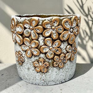 May include: A decorative flower pot with a textured, off-white base and gold-colored floral embellishments. The pot features a cylindrical shape with a slightly ruffled top edge. The flowers are raised and add a three-dimensional effect.