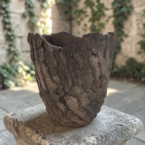 May include: A dark gray, cylindrical vessel with a highly textured, rugged exterior surface that mimics the appearance of tree bark. The rim is uneven and jagged, and the overall form tapers slightly toward the base, creating a deep, open cavity.