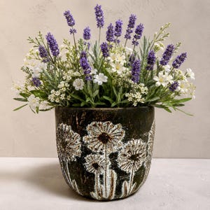 May include: A dark brown, rounded ceramic planter featuring a textured, embossed dandelion pattern in white and gold. The planter is filled with a dense arrangement of purple lavender sprigs and small white flowers with green foliage.