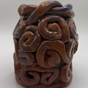 May include: A handmade, brown ceramic vase with a unique, textured design. The vase features a series of swirling, coil-like patterns that create a three-dimensional effect. The top rim has a darker, purplish hue, adding contrast to the overall earthy tones.