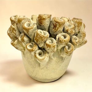 以下が含まれることがあります： ベージュとゴールドの色合いで、多数の丸まった円筒形が特徴的な、ユニークなデザインのセラミック花瓶。 花瓶は丸みを帯びたベースとテクスチャ加工された表面を持ち、有機的で彫刻的な外観を作り出しています。 この装飾品は、センターピースとして使用できます。