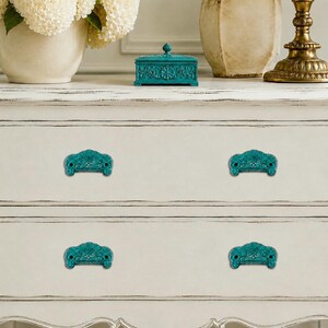 May include: A white wooden dresser with two drawers featuring four ornate teal drawer pulls. Each pull has a detailed, raised floral pattern and a curved shape. The dresser has a distressed finish with visible wood grain and carved edges.