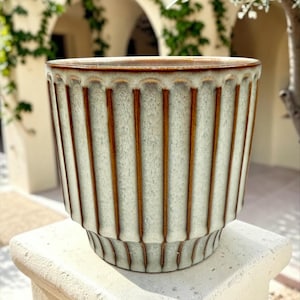 May include: A ceramic planter featuring a vertical ribbed texture with a light cream-colored glaze and contrasting dark brown accents along the ridges and the top rim. The vessel has a tapered base and a wide, open circular top opening.