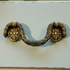May include: A brass-toned metal drawer pull featuring a curved, swinging handle with a leaf-like texture. The mounting plates on each side are oval-shaped and decorated with a raised, central flower motif with radiating petals.