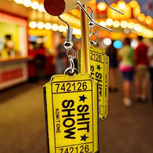 May include: Yellow earrings shaped like vintage show tickets, with the words "SHOW SHIT" and the number "742126" printed on them. The earrings are suspended from silver hooks and are displayed against a blurred background of a carnival.