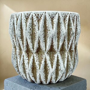 May include: A textured, off-white and brown ceramic planter with a geometric diamond pattern. The planter has a wide, cylindrical shape and sits on a gray, square base. The neutral color palette and textured surface give it a rustic, modern look.