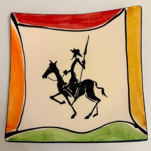 May include: Square ceramic plate with a cream-colored center featuring a black silhouette of a person on horseback holding a spear. The plate has a colorful border with red, orange, yellow, and green sections.