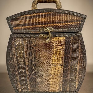 May include: A decorative box with a handle, featuring a reptile skin pattern in shades of brown and gold. The box has a metal clasp and trim, with a curved top and a handle made of a similar material. The overall design suggests a vintage or antique style.