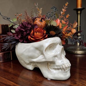 May include: A cream-colored ceramic skull planter filled with an arrangement of dark purple hydrangea, a burnt orange rose, a small orange pumpkin, and various dried autumn-toned foliage including brown leaves and dark berry sprigs.