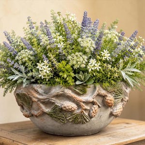 May include: A round, gray stone planter featuring a relief design of pine branches and pinecones around the exterior. The planter is filled with an arrangement of artificial purple lavender, white flowers, and various green foliage.