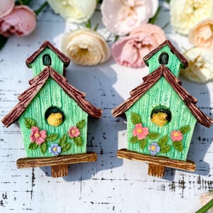 May include: Two identical decorative birdhouses featuring a light green wood-grain texture, a brown roof, and a brown base. Each birdhouse has a central circular opening with a small yellow bird inside, decorated with pink and blue floral accents.