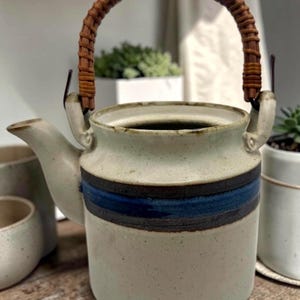 May include: A ceramic teapot with a woven handle. The teapot is off-white with a blue and brown stripe around the middle. The spout is on the left side. The handle is made of woven brown material.