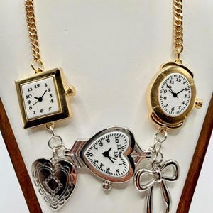 May include: A gold-tone chain necklace featuring multiple watch faces and charms. The necklace includes a square watch, an oval watch, and a heart-shaped watch, all with white faces and black hands. Additional charms include a heart and a bow.