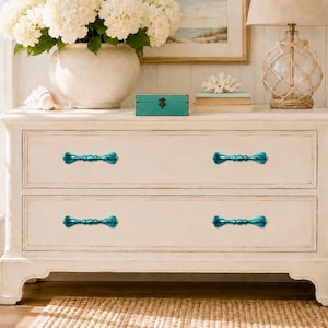 May include: A cream-colored wooden dresser featuring two wide drawers, each equipped with two elongated, metallic teal-colored handles. The dresser has a rectangular frame with a smooth finish and stands on four short, sturdy legs.