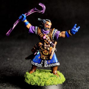 May include: Miniature fantasy figure of a wizard with a staff, wearing blue and purple robes, brown boots, and a leather satchel. The figure is standing on a green, grassy base. The wizard has a beard and is making a hand gesture.