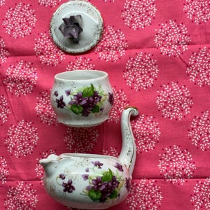 May include: A white porcelain tea set with a floral design featuring purple violets and green leaves. The set includes a teapot with a swan-like spout, a sugar bowl, and a lid with a violet accent. The set is on a pink floral patterned background.