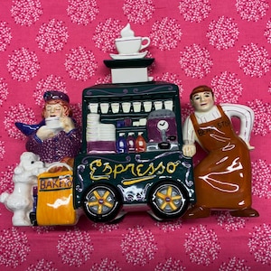 May include: A ceramic espresso cart figurine with a green and gold color scheme. The cart has a sign that says "Espresso" and is flanked by two ceramic figures, one holding a cup and the other wearing an apron that says "Bean Stop".