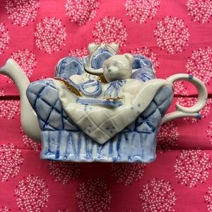 May include: A whimsical teapot in the shape of a blue and white armchair, with a teddy bear and gifts. The teapot has a white spout and handle, and is set against a pink floral background.