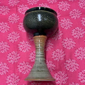 May include: A handmade ceramic chalice with a dark speckled bowl and a tall, flared base. The chalice has a tan, gray, and brown color scheme. The chalice is displayed on a pink background with a white floral pattern.