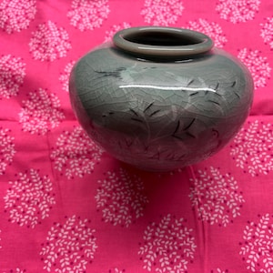 May include: A celadon-colored ceramic vase with a round body and a small opening. The vase features delicate black line drawings of plants and is set against a bright pink background with a white floral pattern. The vase is a decorative item.