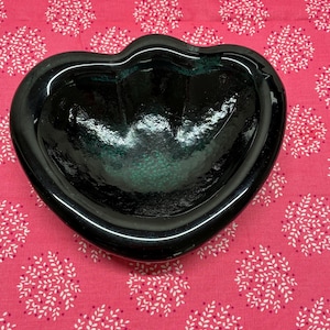 May include: A heart-shaped, dark green glass bowl. The bowl has a glossy finish and a slightly textured interior. The bowl is sitting on a pink fabric with a white floral pattern.