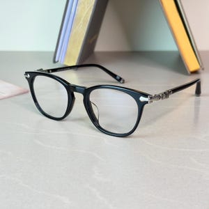 May include: Black-framed eyeglasses with a rounded shape. The temples feature silver decorative accents. The lenses are clear, and the glasses are set against a neutral background. The overall design is modern and stylish.