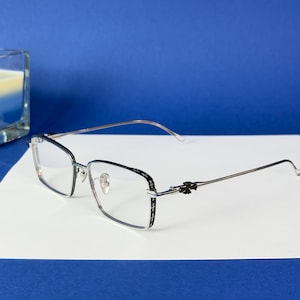May include: Silver-framed eyeglasses with a rectangular shape. The frames have black detailing on the edges and temples, with a decorative cross design on the temple. The lenses are clear, and the glasses are set against a white surface with a blue background.
