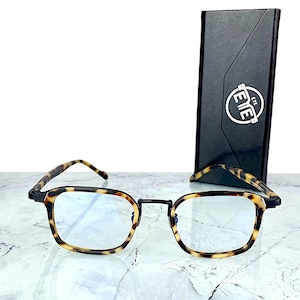 Peut inclure: Lunettes à monture carrée avec un motif écaille de tortue sur les montures et les branches. Les lunettes ont un pont noir et sont accompagnées d'un étui noir avec le mot "EYE" dans un design circulaire.