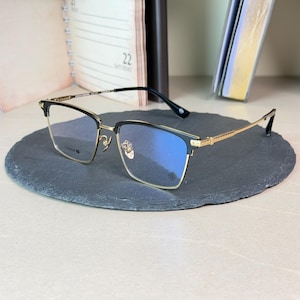 Könnte beinhalten: Goldrandbrille mit schwarzen oberen Rahmen und klaren Gläsern. Die Brille hat eine rechteckige Form und liegt auf einer dunkelgrauen, oval geformten Oberfläche. Die Bügel sind goldfarben und die Gläser haben einen Blaustich.