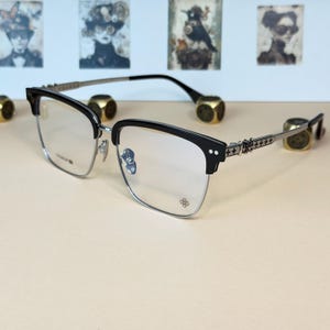 Op de afbeelding: Zwart en zilverkleurige bril met een vierkant montuurontwerp. De bril heeft een zwart bovenframe en zilverkleurige onderframes en -armen. De armen hebben een decoratief kruispatroon. De lenzen zijn helder.