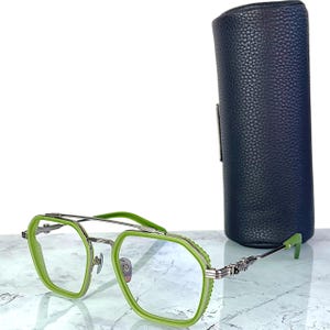 Op de afbeelding: Groene en zilverkleurige bril met een geometrisch montuur. De bril heeft een dubbele brug en wordt geleverd met een donkergroene cilindrische etui. De monturen zijn felgroen, de veren zilver.