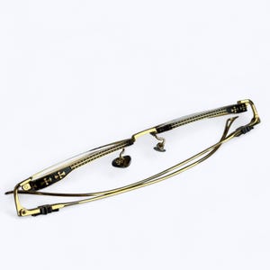 May include: Gold-toned eyeglasses with a rectangular frame and thin temples. The frame features decorative black accents with cross-shaped designs. The lenses are clear, and the nose pads are small and heart-shaped.