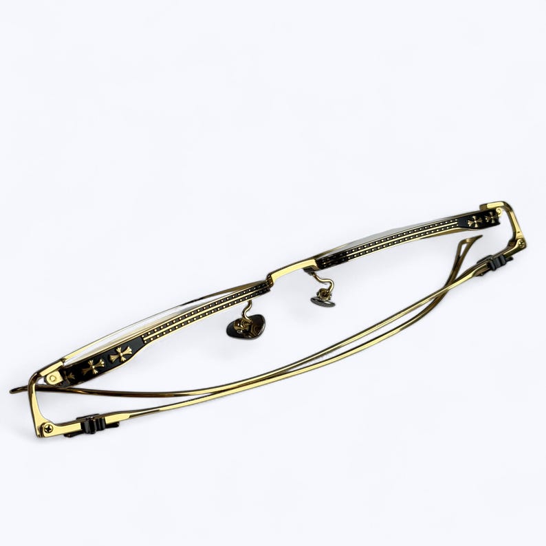 May include: Gold-toned eyeglasses with a rectangular frame and thin temples. The frame features decorative black accents with cross-shaped designs. The lenses are clear, and the nose pads are small and heart-shaped.
