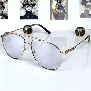Può includere: Occhiali da sole a goccia con sottile montatura in metallo dorato e lenti leggermente colorate. Le aste presentano un motivo tartarugato scuro con la scritta ETE EYEWEAR stampata all'interno. Il ponte ha un design a doppia barra regolabile.