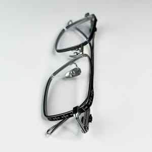 May include: Silver-framed eyeglasses with a black cross pattern on the temples. The glasses have a rectangular shape and clear lenses. The nose pads are silver and the arms are thin and curved.