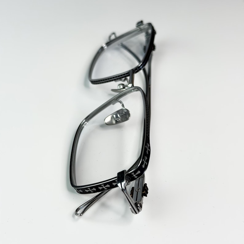 May include: Silver-framed eyeglasses with a black cross pattern on the temples. The glasses have a rectangular shape and clear lenses. The nose pads are silver and the arms are thin and curved.