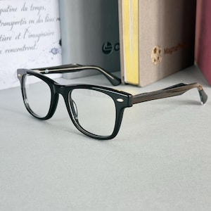 Op de afbeelding: Zwarte bril met een rechthoekig montuur en heldere glazen. De pootjes zijn van donker hout. De bril ligt op een lichtgrijze ondergrond, met boeken op de achtergrond.