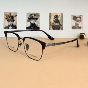 May include: Eyeglasses with a dark brown and silver frame. The lenses are clear, and the arms have a decorative design. The glasses have a titanium label. The text "WENZIKEUOXIN" is visible on the frame.