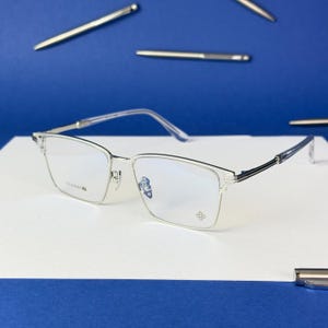 Pode incluir: Óculos com armação prateada de formato retangular. Os óculos possuem lentes transparentes e um design delicado, com o texto "TITANIUM" visível na armação. O fundo é um gradiente de branco e azul, com várias canetas prateadas espalhadas.