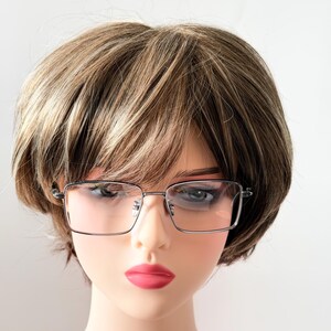 May include: Silver-framed eyeglasses with rectangular lenses are displayed on a mannequin head with a wig. The wig is a short, layered style with brown and blonde highlights. The mannequin has pink lipstick.