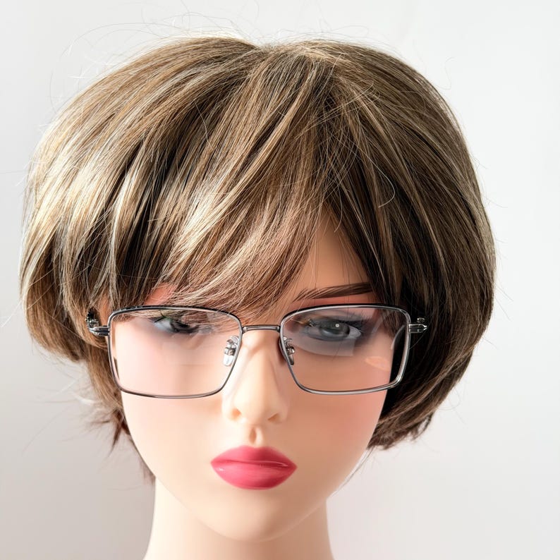 May include: Silver-framed eyeglasses with rectangular lenses are displayed on a mannequin head with a wig. The wig is a short, layered style with brown and blonde highlights. The mannequin has pink lipstick.