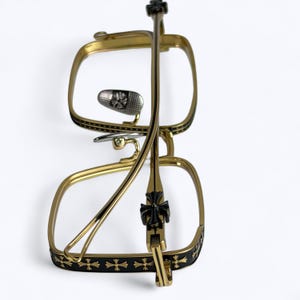 May include: Gold-toned eyeglasses with a square frame and black accents. The frames feature a decorative cross pattern and a silver nose bridge. The temples are thin and gold, with a black cross detail near the hinge.