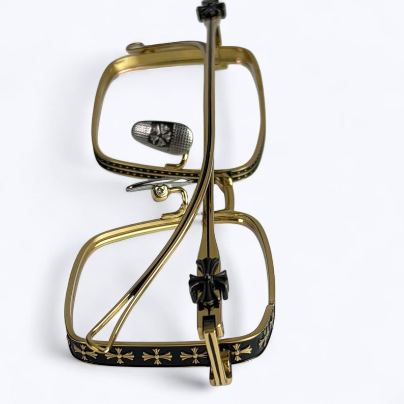 May include: Gold-toned eyeglasses with a square frame and black accents. The frames feature a decorative cross pattern and a silver nose bridge. The temples are thin and gold, with a black cross detail near the hinge.